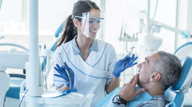 A female dentist smiling at her male patient while checking his teeth as the patient gently holds his mouth